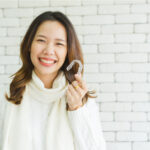 Asian woman in a white turtleneck sweater smiles while holding her Invisalign aligner