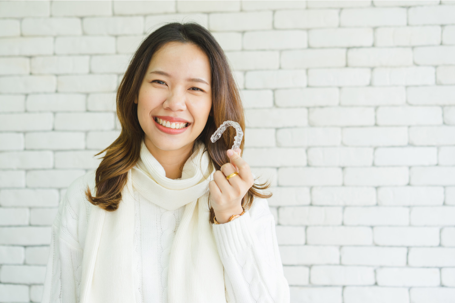 Asian woman in a white turtleneck sweater smiles while holding her Invisalign aligner